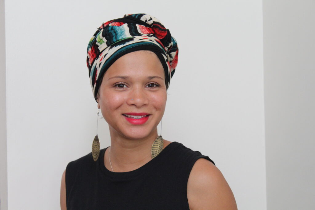 A portrait of a smiling Black woman wearing red lipstick, dangling earrings, and a patterned hair wrap