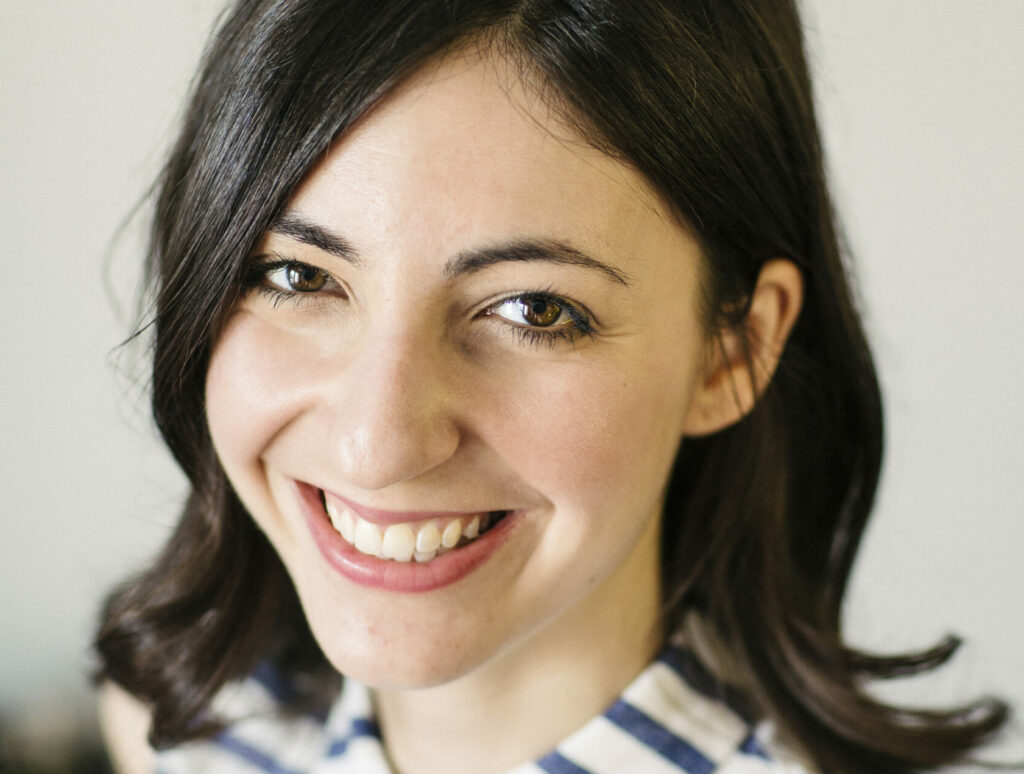 A white woman with black hair smiling against a blurred gray background