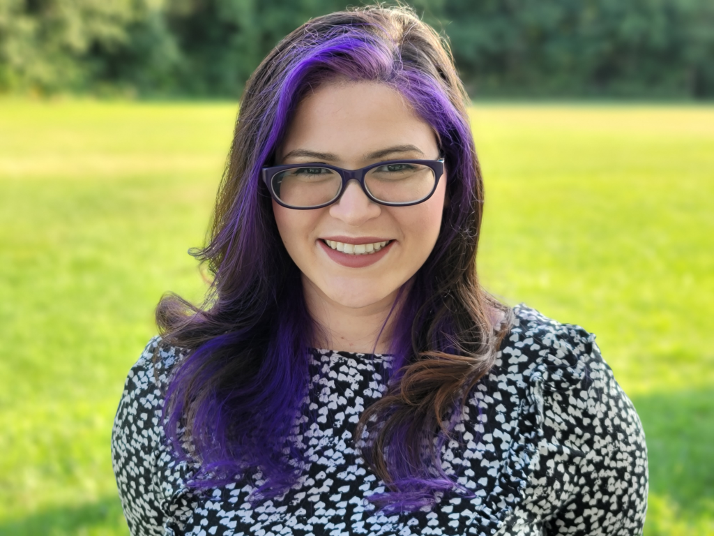 A smiling woman with purple hair and glasses against a natural background