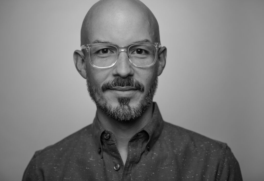 Black and white photo of Antonio Garcia who has a shaved head, thick rimmed glasses, mustache and beard, with a collared shirt.